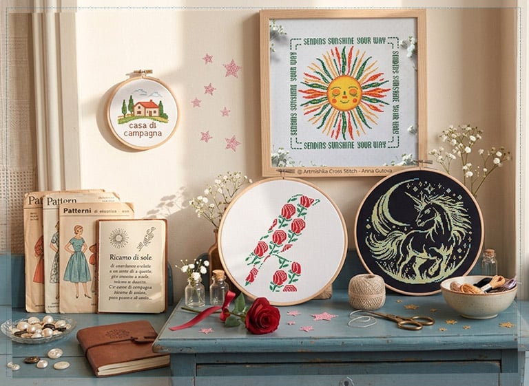 Ricami a punto croce Artmishka su un tavolo vintage: sole sorridente, iniziale floreale e unicorno, tra cartamodelli e fiori di campo.